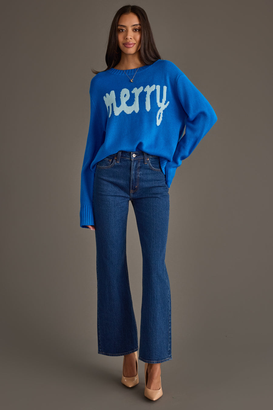 A model wears a Panache Apparel Collection Royal Blue & Light Blue Merry Sweater with light blue script on the front, paired with dark wash flared jeans and beige heels.