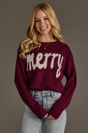 A smiling woman wears a Wine Merry Sweater in burgundy with the word merry in white text across the chest, paired with light-wash blue jeans.