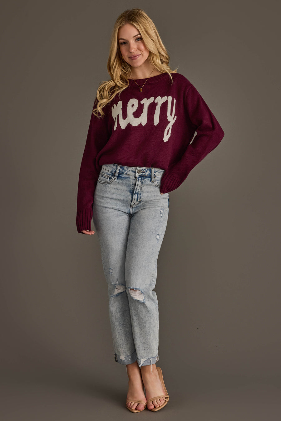 A person wearing a Wine Merry Sweater in maroon with the word merry in white letters, paired with light-wash distressed jeans and beige heels.