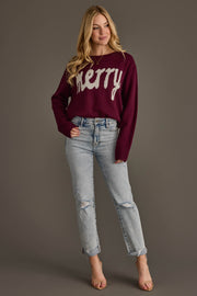 A woman wears a Panache Apparel Collection Wine Merry Sweater with white lettering across the chest, paired with light-wash distressed jeans.