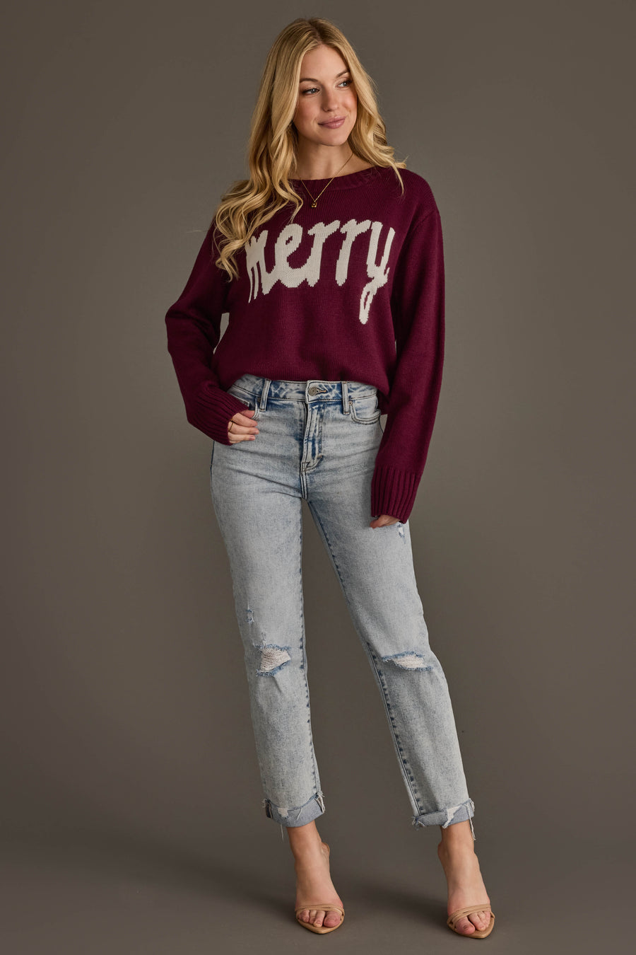 A woman wears a Panache Apparel Collection Wine Merry Sweater with white lettering across the chest, paired with light-wash distressed jeans.