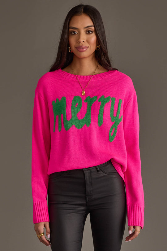 A model wears a Bright Pink & Green Merry Sweater, a bright pink long-sleeved sweater with the word merry written in large green letters across the front.