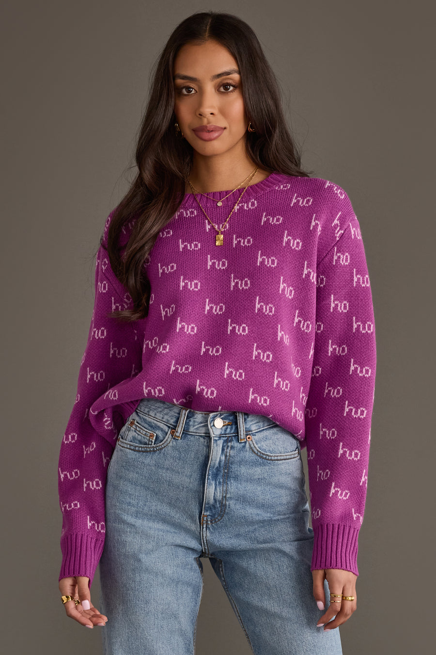A woman wearing a purple long-sleeved Purple Ho Ho Ho Sweater with a white repeating pattern, paired with light-wash blue jeans.