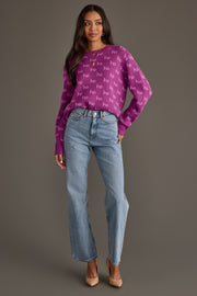 A woman wearing a Purple Ho Ho Ho Sweater with a repeating white pattern, paired with light-wash straight-leg jeans and neutral heels.