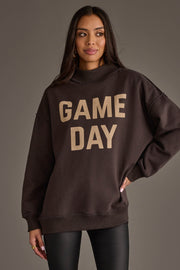 A woman models the Panache Apparel Collection’s Dark Gray Game Day & Sunday Funday Reversible Sweatshirt, featuring bold beige "GAME DAY" lettering on the front, paired with black pants, against a plain gray backdrop.