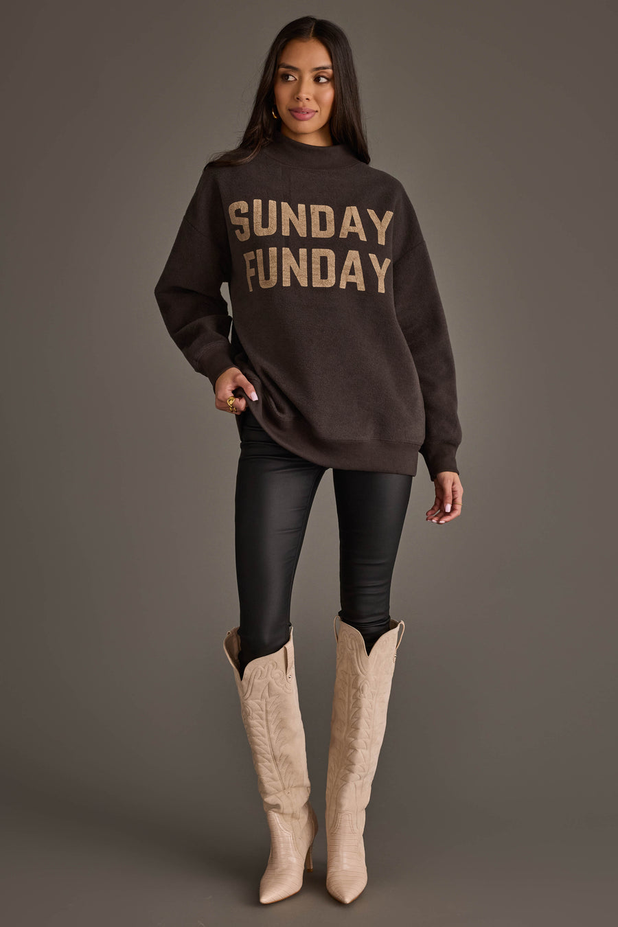 A woman stands against a gray background wearing the Panache Apparel Collection Dark Gray Game Day & Sunday Funday Reversible Sweatshirt, paired with black leggings and tall beige cowboy boots; she has long dark hair and strikes a relaxed pose.