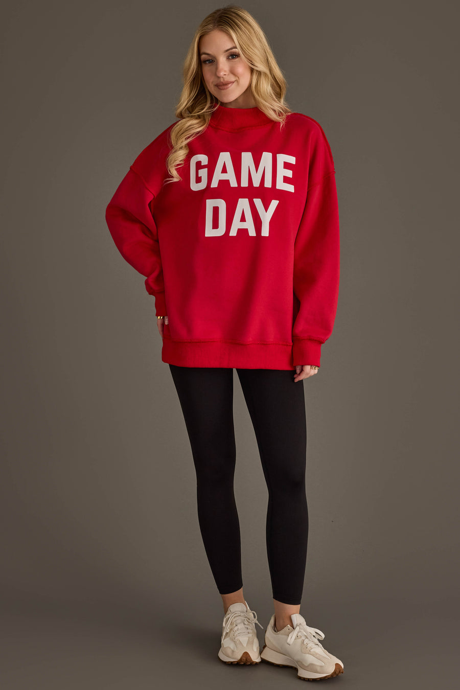 A woman stands against a plain background, smiling with her hands at her sides. She wears the Panache Apparel Collection Red Game Day & Sunday Funday Reversible Sweatshirt, paired with black leggings and white sneakers.