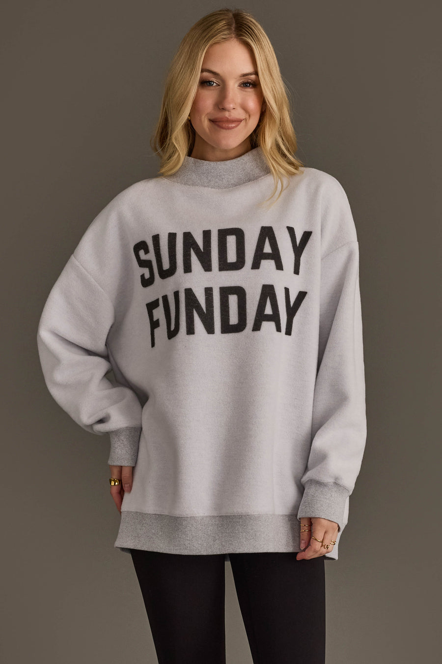 A woman with long blond hair wears the Panache Apparel Collection Light Gray Game Day & Sunday Funday Reversible Sweatshirt, styled with black leggings, standing against a plain, dark background.