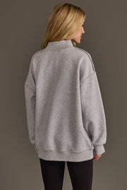 A person with long blonde hair, wearing the Panache Apparel Collection Light Gray Game Day & Sunday Funday Reversible Sweatshirt and black pants, stands facing away against a plain brown background.