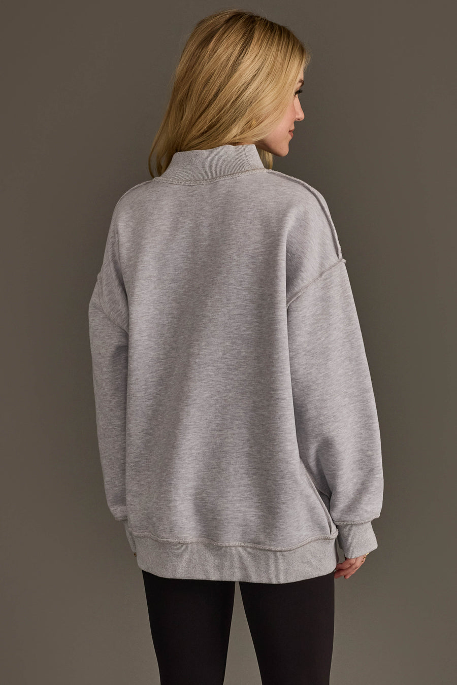 A person with long blonde hair, wearing the Panache Apparel Collection Light Gray Game Day & Sunday Funday Reversible Sweatshirt and black pants, stands facing away against a plain brown background.
