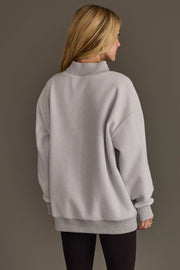 A woman with long blonde hair stands facing away, wearing the Panache Apparel Collection Light Gray Game Day & Sunday Funday Reversible Sweatshirt and black leggings against a plain, neutral background.