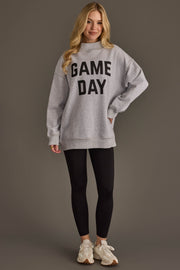 A woman stands against a plain background wearing the Panache Apparel Collection Light Gray Game Day & Sunday Funday Reversible Sweatshirt, paired with black leggings and white sneakers, with one hand on her hip and long, wavy blonde hair.