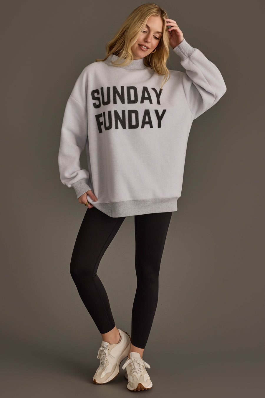 A woman stands smiling against a plain background, wearing the Panache Apparel Collection Light Gray Game Day & Sunday Funday Reversible Sweatshirt, paired with black leggings and white sneakers.