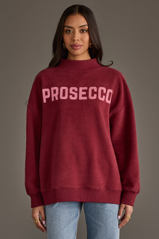 A person wears a Burgundy Wine & Prosecco Reversible Sweatshirt featuring a burgundy mock-neck with the word PROSECCO printed in large pink letters across the chest.