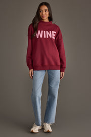 A person wearing a burgundy Burgundy Wine & Prosecco Reversible Sweatshirt with the word WINE in light pink lettering, paired with blue jeans and sneakers.