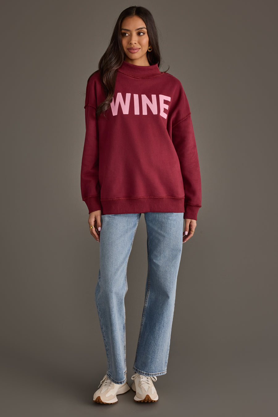 A person wearing a burgundy Burgundy Wine & Prosecco Reversible Sweatshirt with the word WINE in light pink lettering, paired with blue jeans and sneakers.