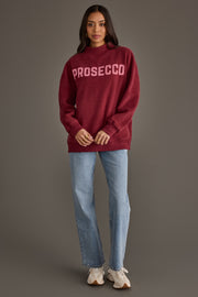 A person wearing a burgundy Burgundy Wine & Prosecco Reversible Sweatshirt with PROSECCO printed on the front, light-wash jeans, and athletic sneakers.