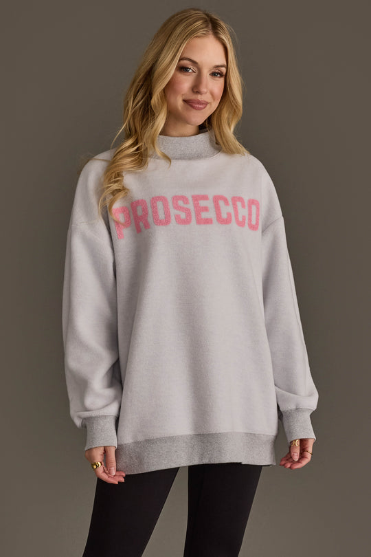 A woman with long blonde hair wears the Panache Apparel Collection Light Gray Wine & Prosecco Reversible Sweatshirt paired with black pants, standing against a plain gray background.