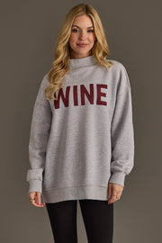 A woman with long blonde hair is seen wearing the Panache Apparel Collection’s Light Gray Wine & Prosecco Reversible Sweatshirt, featuring the word WINE in bold red letters on the front, paired with black leggings as she stands against a neutral background.