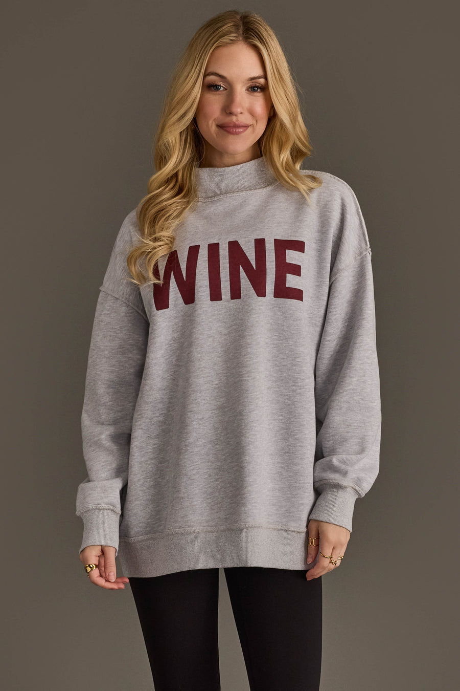 A woman with long blonde hair is seen wearing the Panache Apparel Collection’s Light Gray Wine & Prosecco Reversible Sweatshirt, featuring the word WINE in bold red letters on the front, paired with black leggings as she stands against a neutral background.