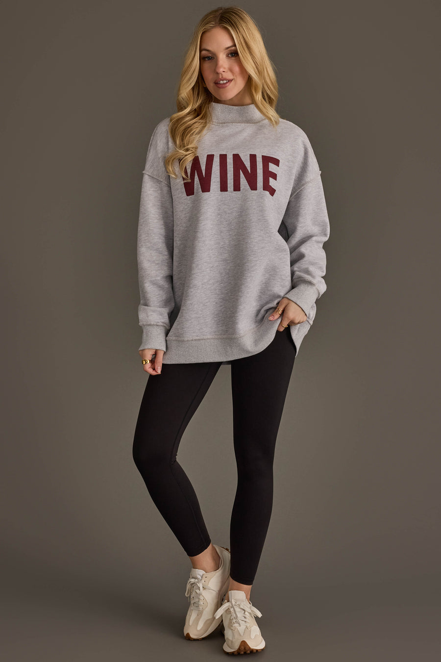 A woman with long blonde hair stands and poses against a plain brown background, wearing the Panache Apparel Collection Light Gray Wine & Prosecco Reversible Sweatshirt, paired with black leggings and beige sneakers.