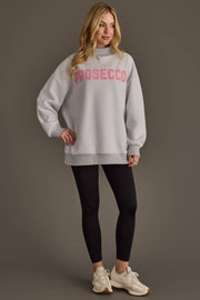 A woman stands smiling against a plain gray background, wearing the Panache Apparel Collection Light Gray Wine & Prosecco Reversible Sweatshirt with pink lettering, paired with black leggings and white sneakers.