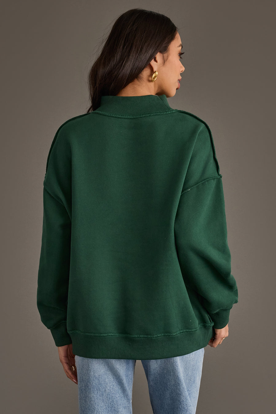 Back view of a person wearing a dark green Dark Green Nice & Naughty Reversible Sweatshirt with a mock neckline and dropped shoulders, paired with light wash blue jeans.