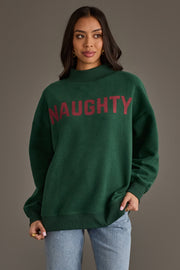 A woman wearing a Dark Green Nice & Naughty Reversible Sweatshirt, featuring a dark green mock-neck design with the word NAUGHTY printed in large red letters across the chest.