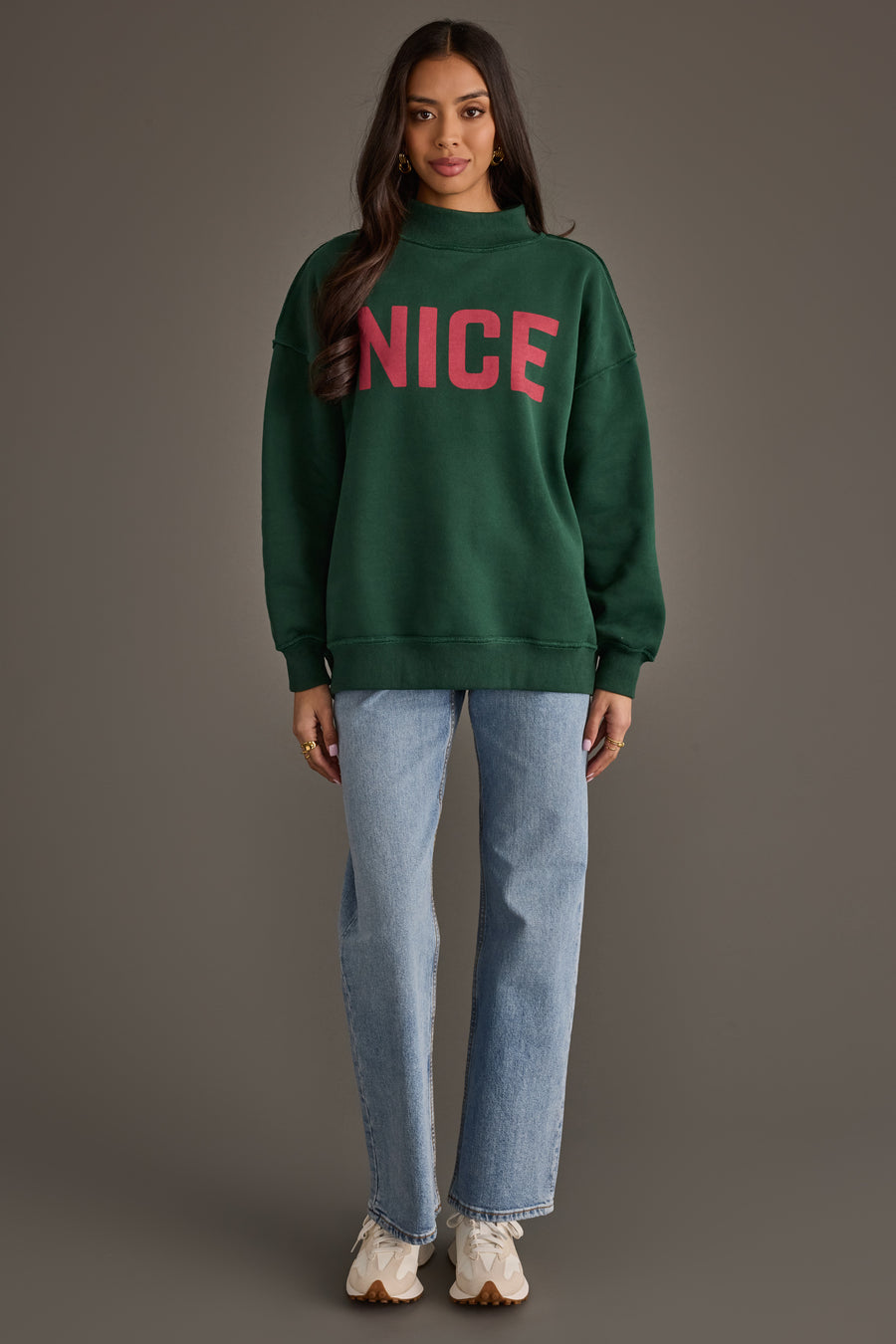A person wearing a Dark Green Nice & Naughty Reversible Sweatshirt with the word NICE in large red letters, light-wash jeans, and white sneakers.
