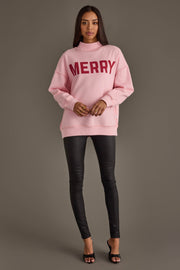 A person wearing a Pink Merry & Cheers Reversible Sweatshirt in light pink with the word MERRY printed in dark red across the chest, paired with black fitted pants.