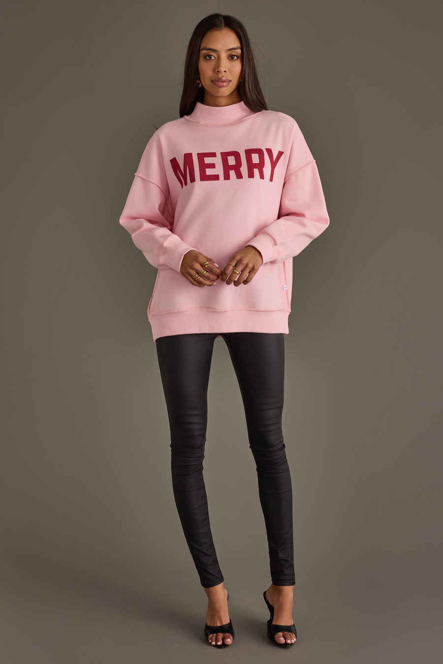 A person wearing a Pink Merry & Cheers Reversible Sweatshirt in light pink with the word MERRY printed in dark red across the chest, paired with black fitted pants.