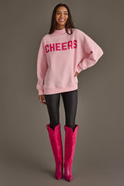 A smiling woman wears a pink long-sleeve Pink Merry & Cheers Reversible Sweatshirt with the word CHEERS in red letters, paired with black pants and bright pink knee-high boots.