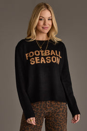 A woman with long blonde hair is wearing the Panache Apparel Collection's Black Football Season Sweater, featuring "FOOTBALL SEASON" in tan text, paired with leopard print pants. She stands against a plain gray background, smiling at the camera.
