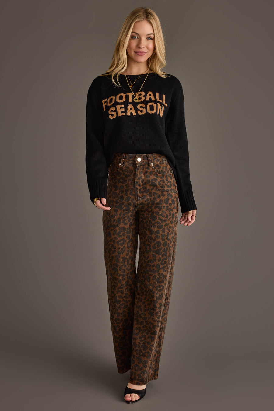 A woman stands smiling in the Panache Apparel Collection's Black Football Season Sweater, featuring "FOOTBALL SEASON" in orange letters, paired with brown wide-legged leopard print pants as she poses against a plain gray background.