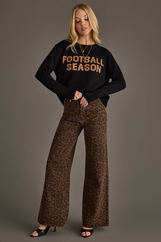 A woman poses against a plain gray background wearing the Panache Apparel Collection Black Football Season Sweater, styled with brown leopard print wide-leg pants and black open-toe heels.