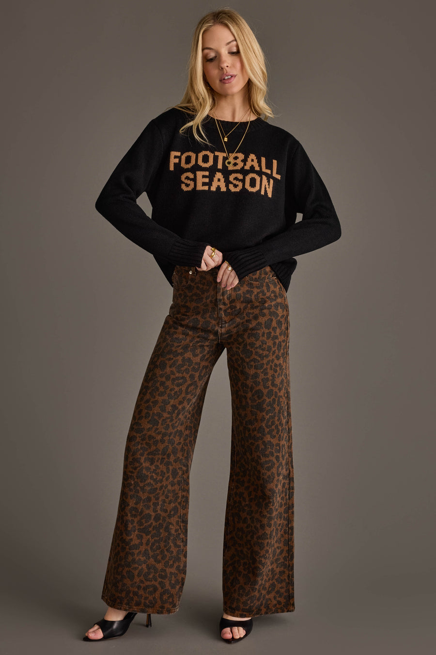 A woman poses against a plain gray background wearing the Panache Apparel Collection Black Football Season Sweater, styled with brown leopard print wide-leg pants and black open-toe heels.