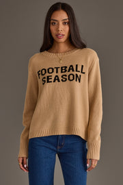 A woman with long dark hair wears the Panache Apparel Collection’s Tan Football Season Sweater, featuring black "FOOTBALL SEASON" lettering, paired with blue jeans and standing against a plain gray background.