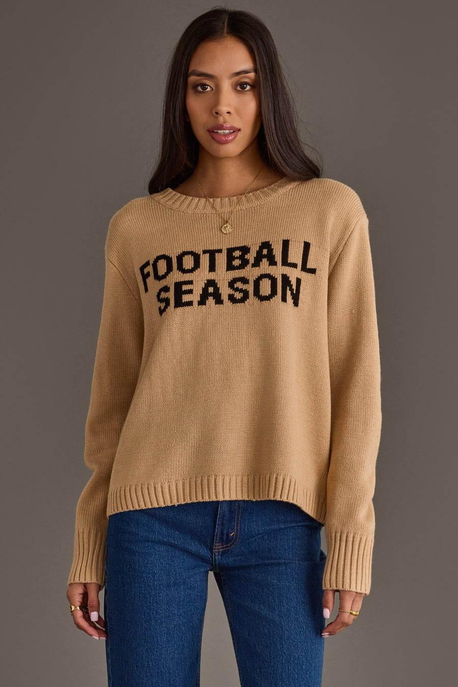 A woman with long dark hair wears the Panache Apparel Collection’s Tan Football Season Sweater, featuring black "FOOTBALL SEASON" lettering, paired with blue jeans and standing against a plain gray background.