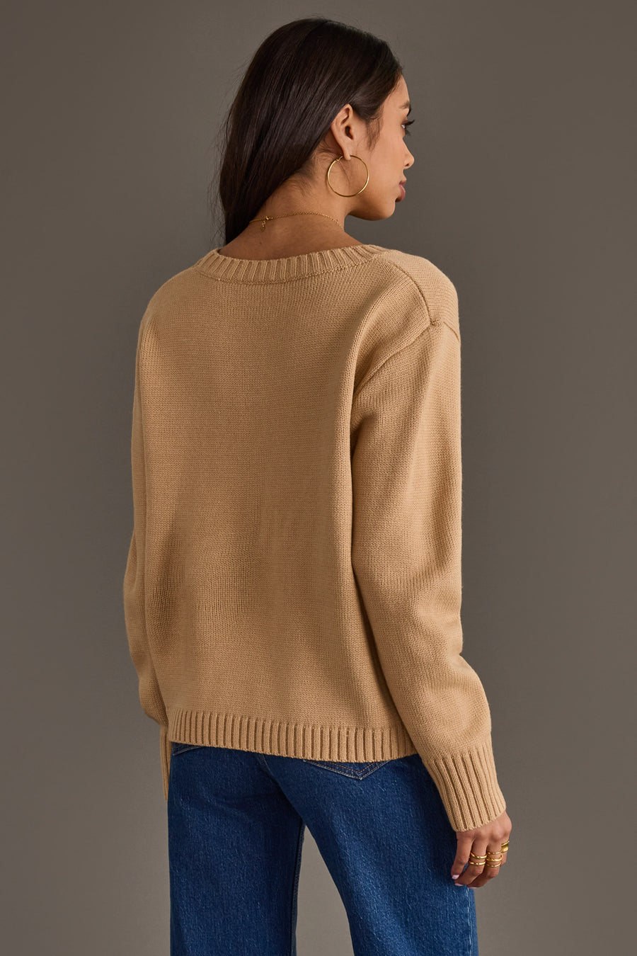 Wearing the Tan Football Season Sweater from Panache Apparel Collection, a woman with long dark hair, gold hoop earrings, and blue jeans stands facing away against a plain gray background.
