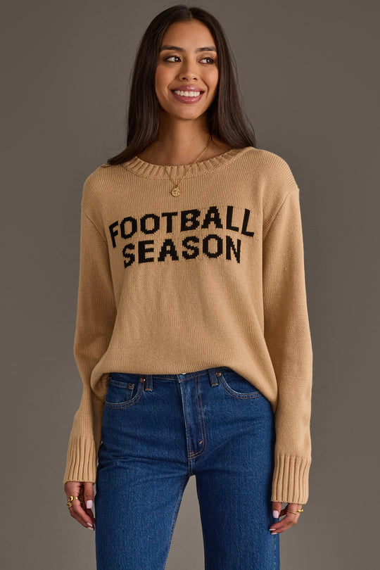 A woman models the Tan Football Season Sweater from Panache Apparel Collection, featuring “FOOTBALL SEASON” in bold black letters. She stands smiling with relaxed hands, pairing the sweater with blue jeans against a plain gray background.