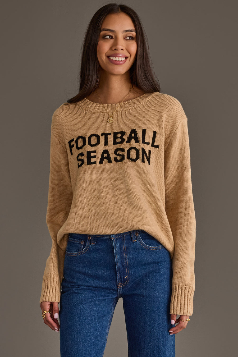 A woman models the Tan Football Season Sweater from Panache Apparel Collection, featuring “FOOTBALL SEASON” in bold black letters. She stands smiling with relaxed hands, pairing the sweater with blue jeans against a plain gray background.