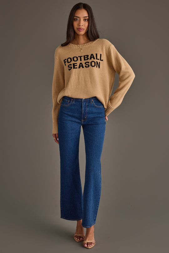A woman stands against a plain background wearing the Panache Apparel Collection’s Tan Football Season Sweater, paired with blue jeans and heeled sandals. She has long hair and gazes at the camera with a relaxed expression.
