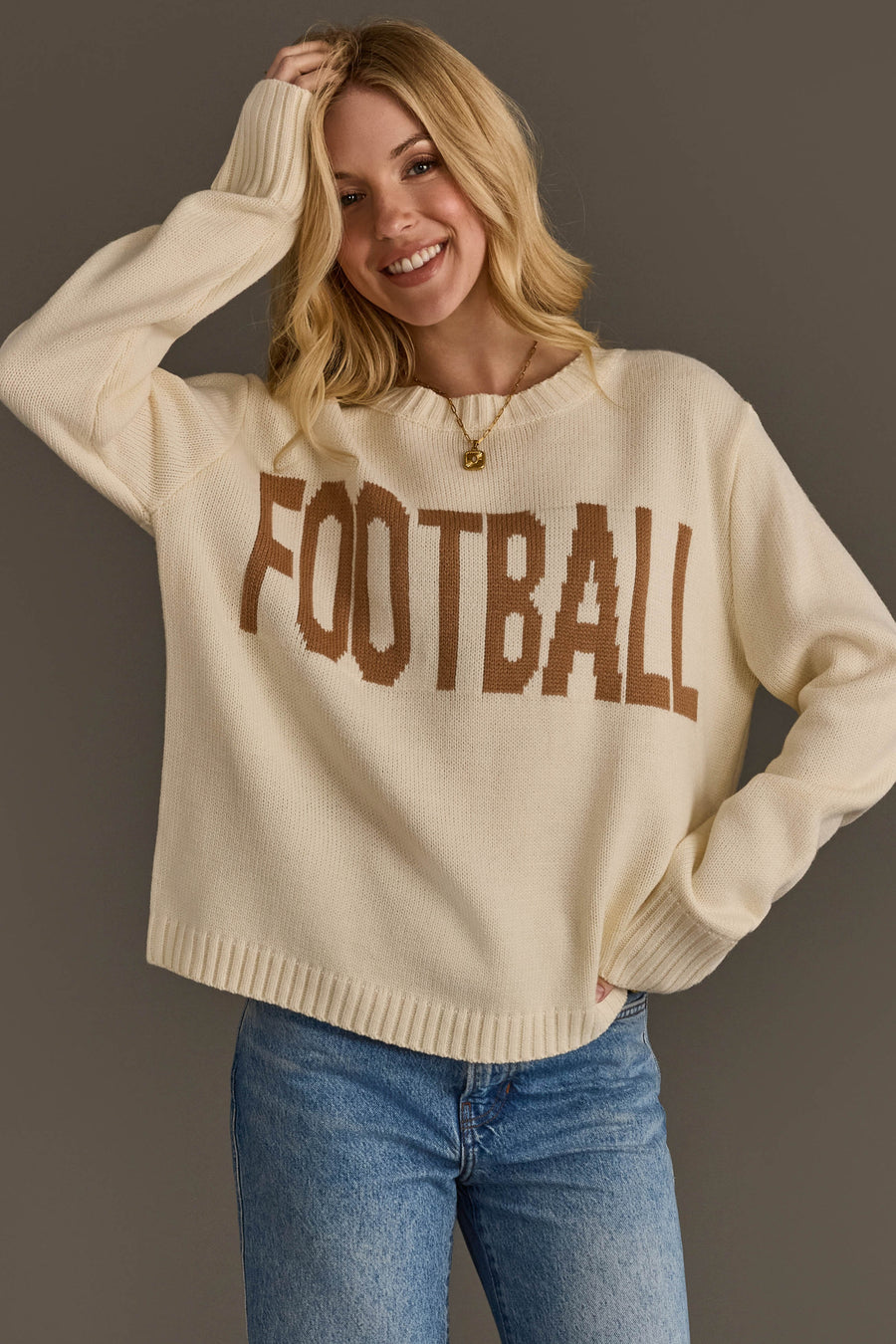 A person wearing the Panache Apparel Collection’s Ivory Football Sweater, featuring the word “FOOTBALL” in bold brown letters, paired with blue jeans, stands against a plain gray background, smiling with one hand on their hip and the other touching their head.