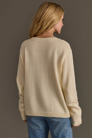 A woman with blonde hair, dressed in the Panache Apparel Collection Ivory Football Sweater and blue jeans, faces away from the camera against a plain, neutral background.
