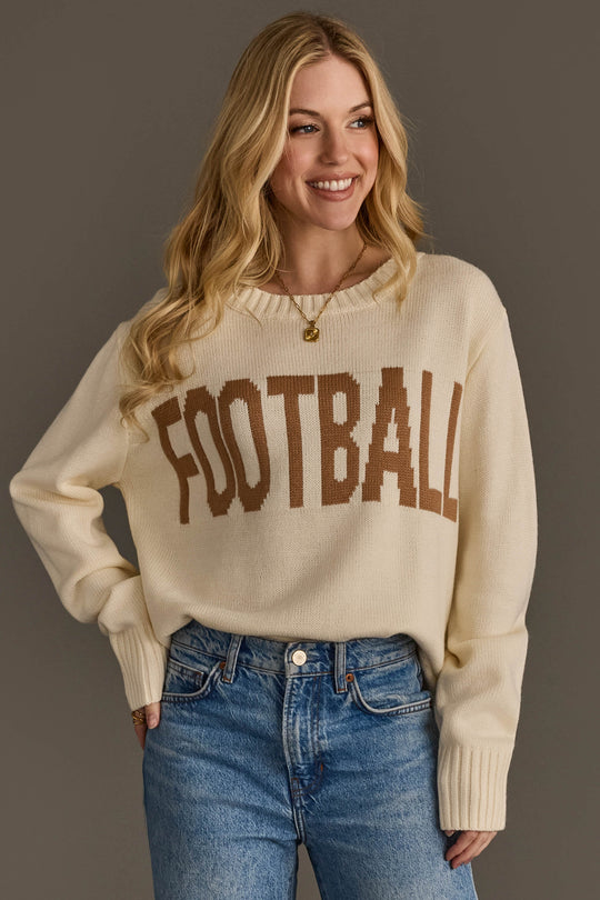 A smiling person wears the Panache Apparel Collection's Ivory Football Sweater, featuring "FOOTBALL" in brown lettering, paired with blue jeans and standing against a plain gray background.