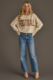 A smiling person with long blonde hair wears the Ivory Football Sweater by Panache Apparel Collection, featuring "FOOTBALL" in brown letters, paired with blue jeans and beige heels, standing against a plain brown background.