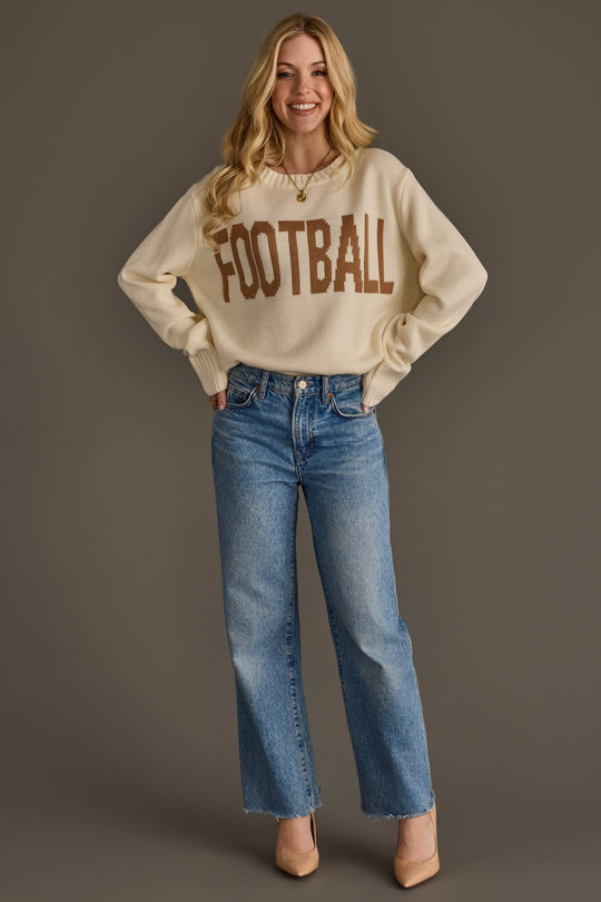 A smiling person with long blonde hair wears the Ivory Football Sweater by Panache Apparel Collection, featuring "FOOTBALL" in brown letters, paired with blue jeans and beige heels, standing against a plain brown background.