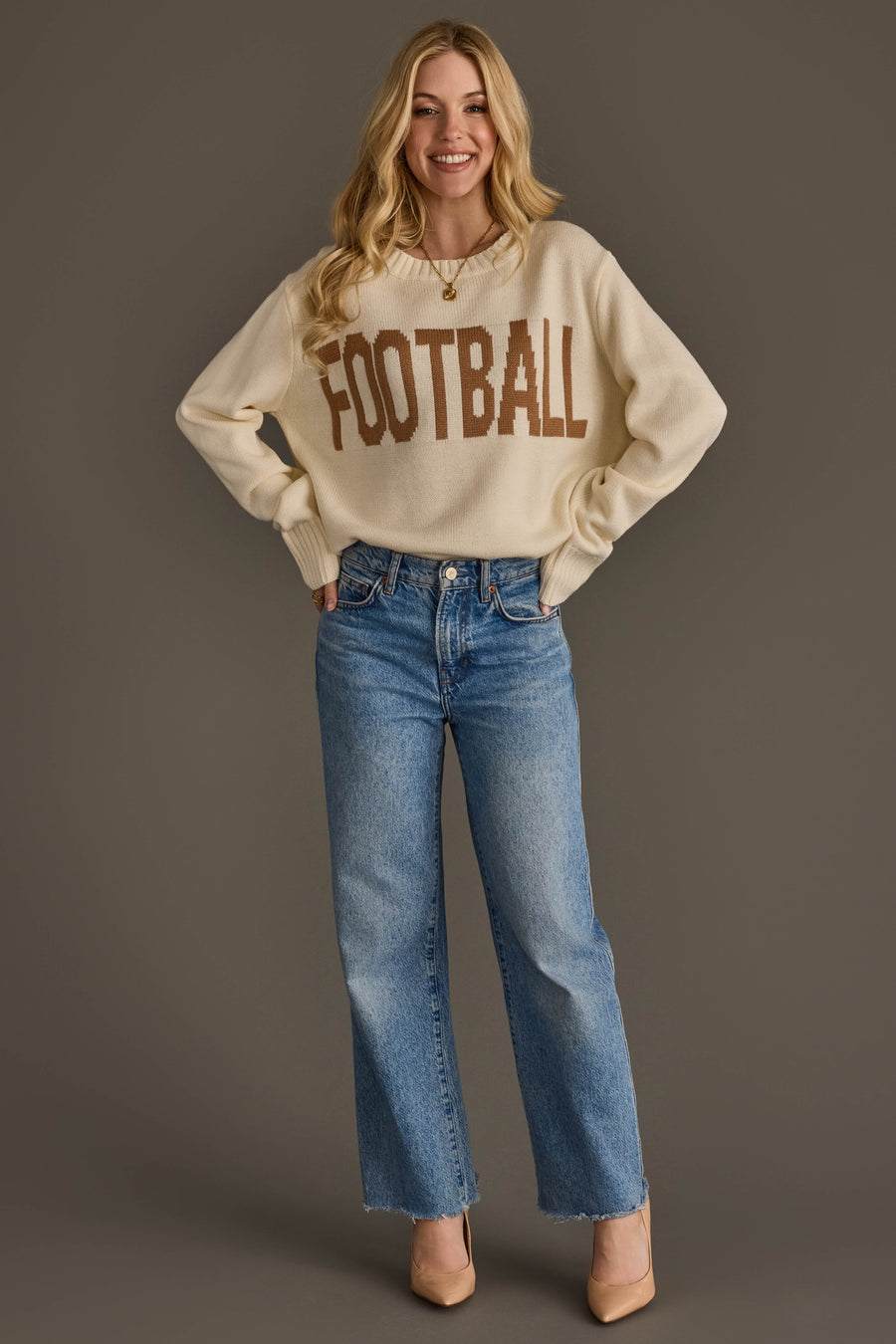 A smiling person with long blonde hair wears the Ivory Football Sweater by Panache Apparel Collection, featuring "FOOTBALL" in brown letters, paired with blue jeans and beige heels, standing against a plain brown background.