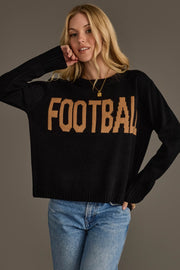 A person with long blonde hair models the Black Football Sweater from Panache Apparel Collection—featuring "FOOTBALL" in tan letters—paired with blue jeans and posing with one arm bent and a relaxed expression against a plain background.