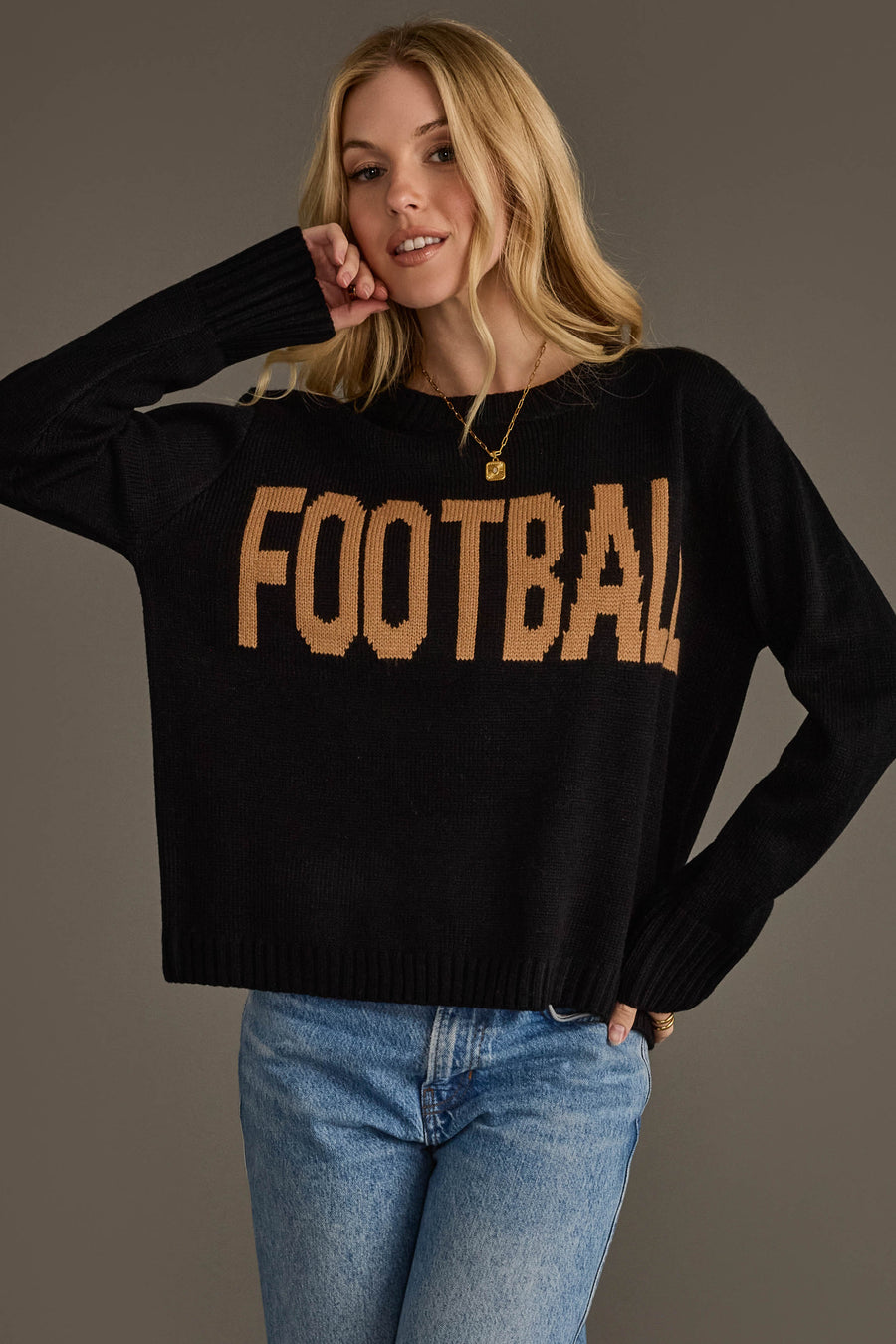 A person with long blonde hair models the Black Football Sweater from Panache Apparel Collection—featuring "FOOTBALL" in tan letters—paired with blue jeans and posing with one arm bent and a relaxed expression against a plain background.